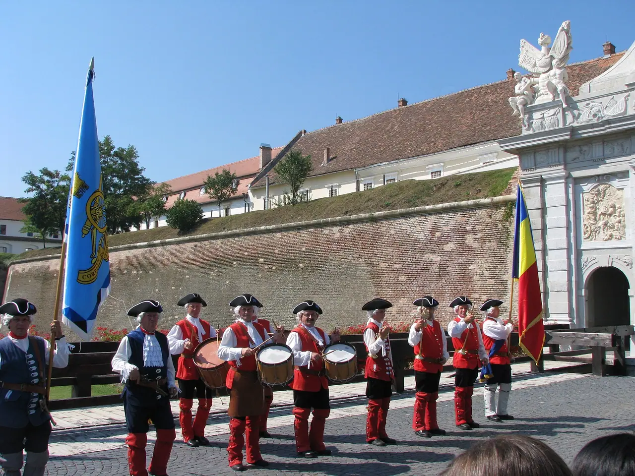 Fortress guards in red and white eighteenth-century uniforms with tricorne hats and drums lined up on the cobbles below the brick rampart and the carved stone gate at Alba Carolina