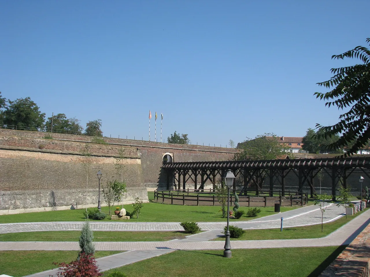 A long covered timber gallery running along the landscaped outer ditch of Alba Carolina, below the brick rampart with flags on top