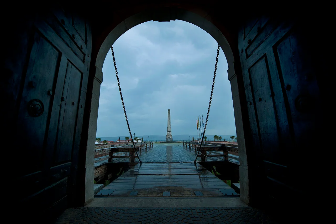 View out through the open studded wooden doors of a gate at Alba Carolina, along the chains and timber deck of the drawbridge towards the tall stone obelisk on the esplanade