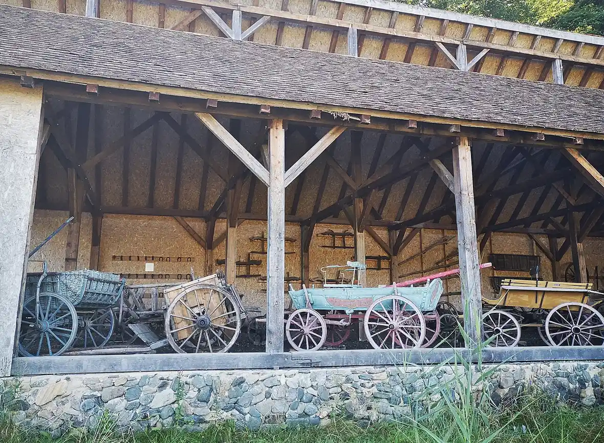 A row of horse-drawn carts and wagons under a wood-shingled shed roof at ASTRA Museum, tools and spare wheels along the back wall
