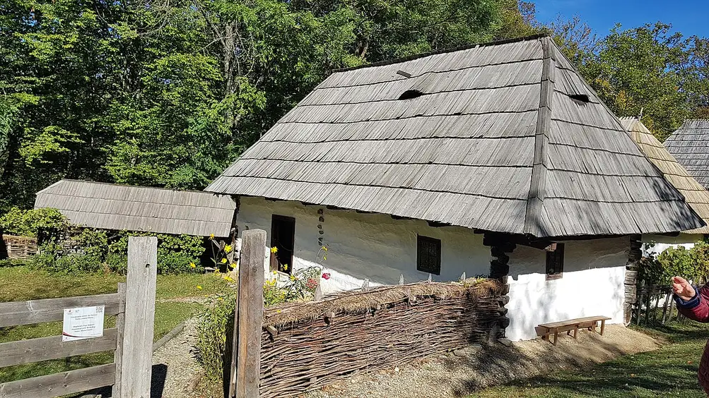 A whitewashed farmhouse with a steep wood-shingled roof behind a woven wattle fence at ASTRA Museum