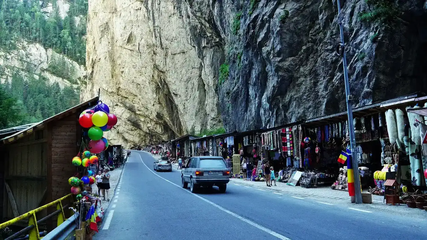 National road DN12C running along the floor of the Bicaz Gorge between limestone walls, with a line of wooden souvenir kiosks on the right and a car coming towards the camera