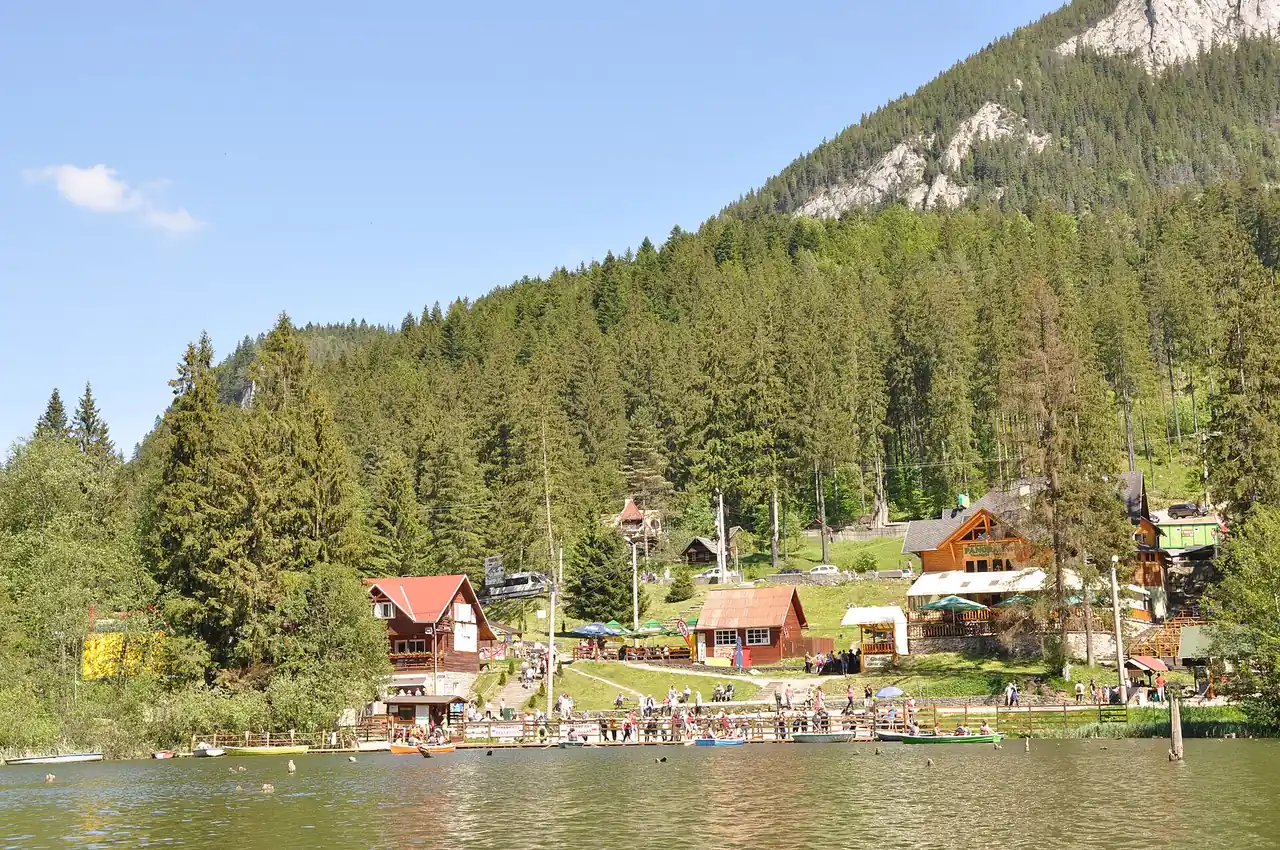 The shore of Lacu Rosu seen from a boat, with wooden pensions, terraces and kiosks along the waterline and a forested ridge rising behind them