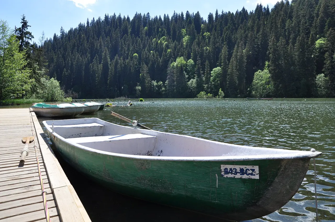 A green wooden rowing boat tied to a timber jetty on Lacu Rosu, with more boats moored beyond and dense spruce forest on the far shore