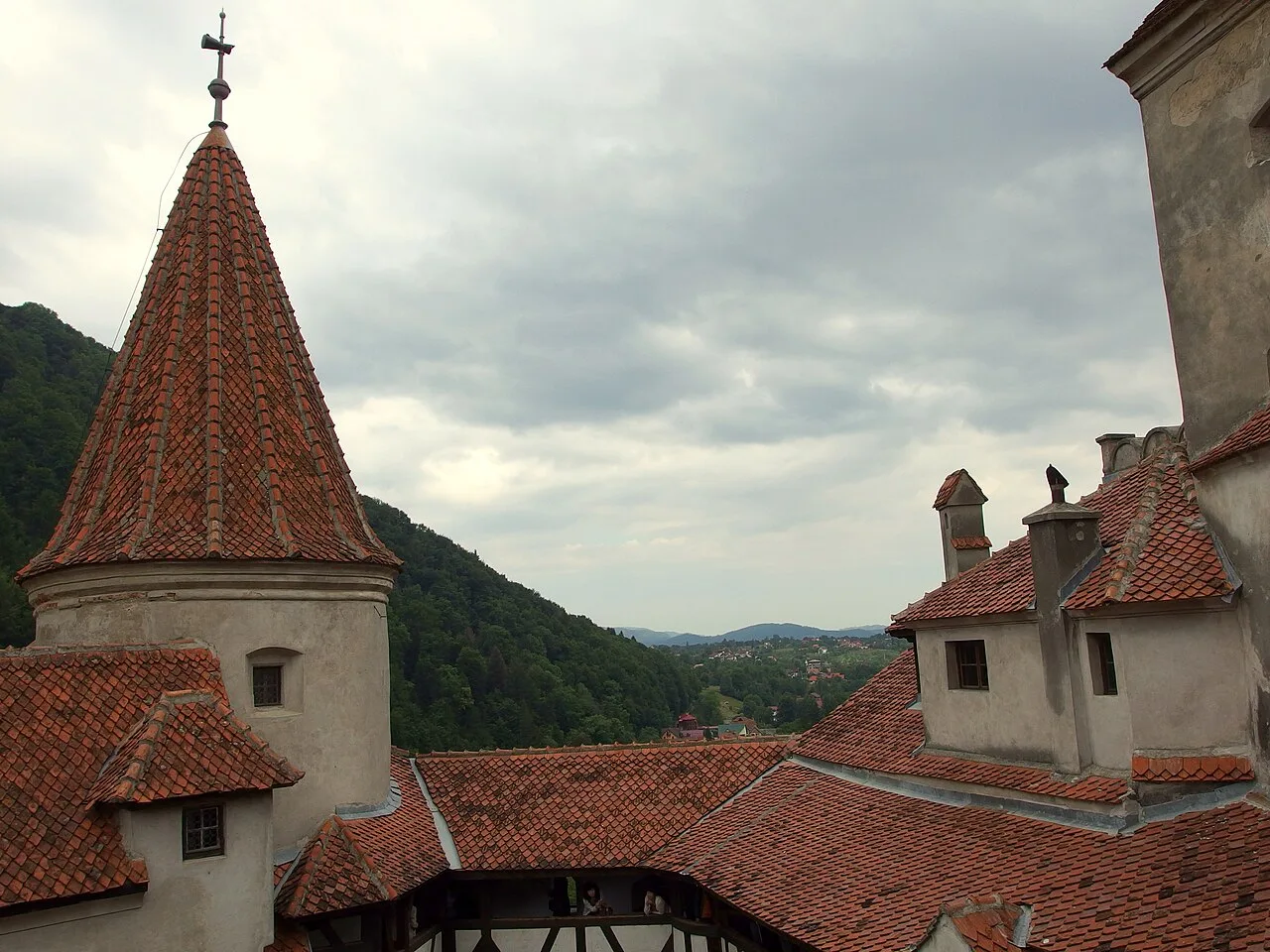 The red-tiled turret and roofs of Bran Castle in the foreground with wooded hills and a village in the valley beyond under a grey sky