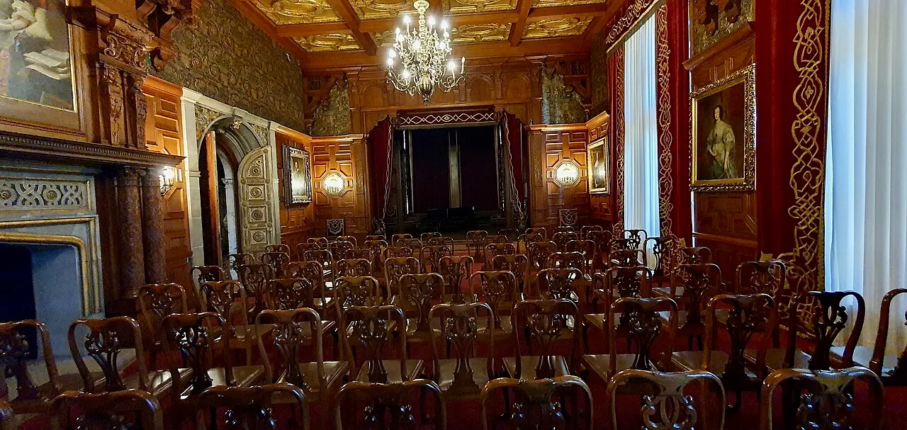 A wood-panelled hall inside Peles Castle with carved walnut walls, a chandelier, gilt-framed portraits and rows of chairs