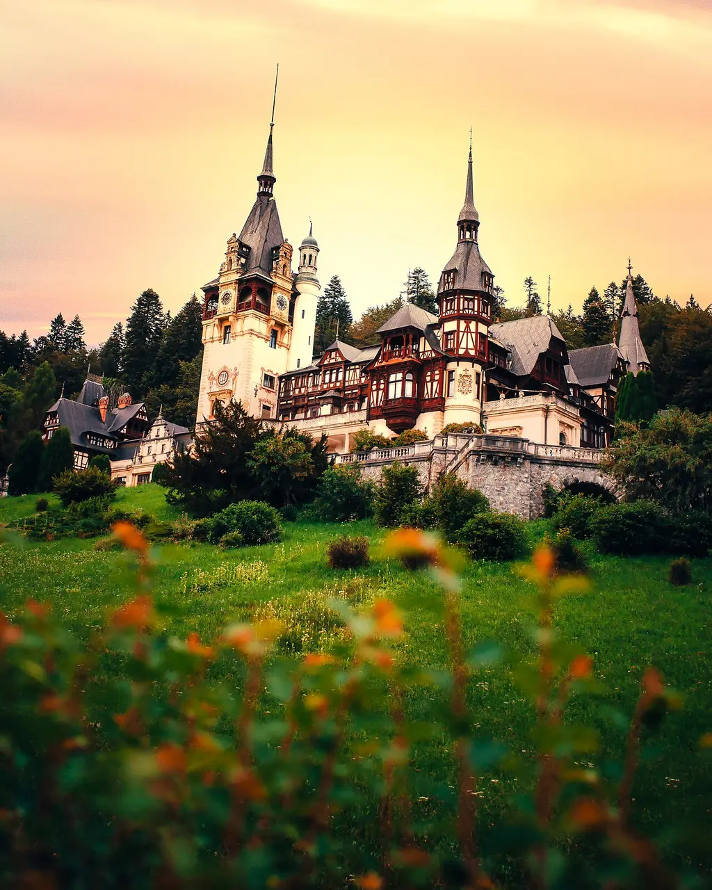 Peles Castle at sunset, its towers glowing against an orange sky above a wildflower meadow