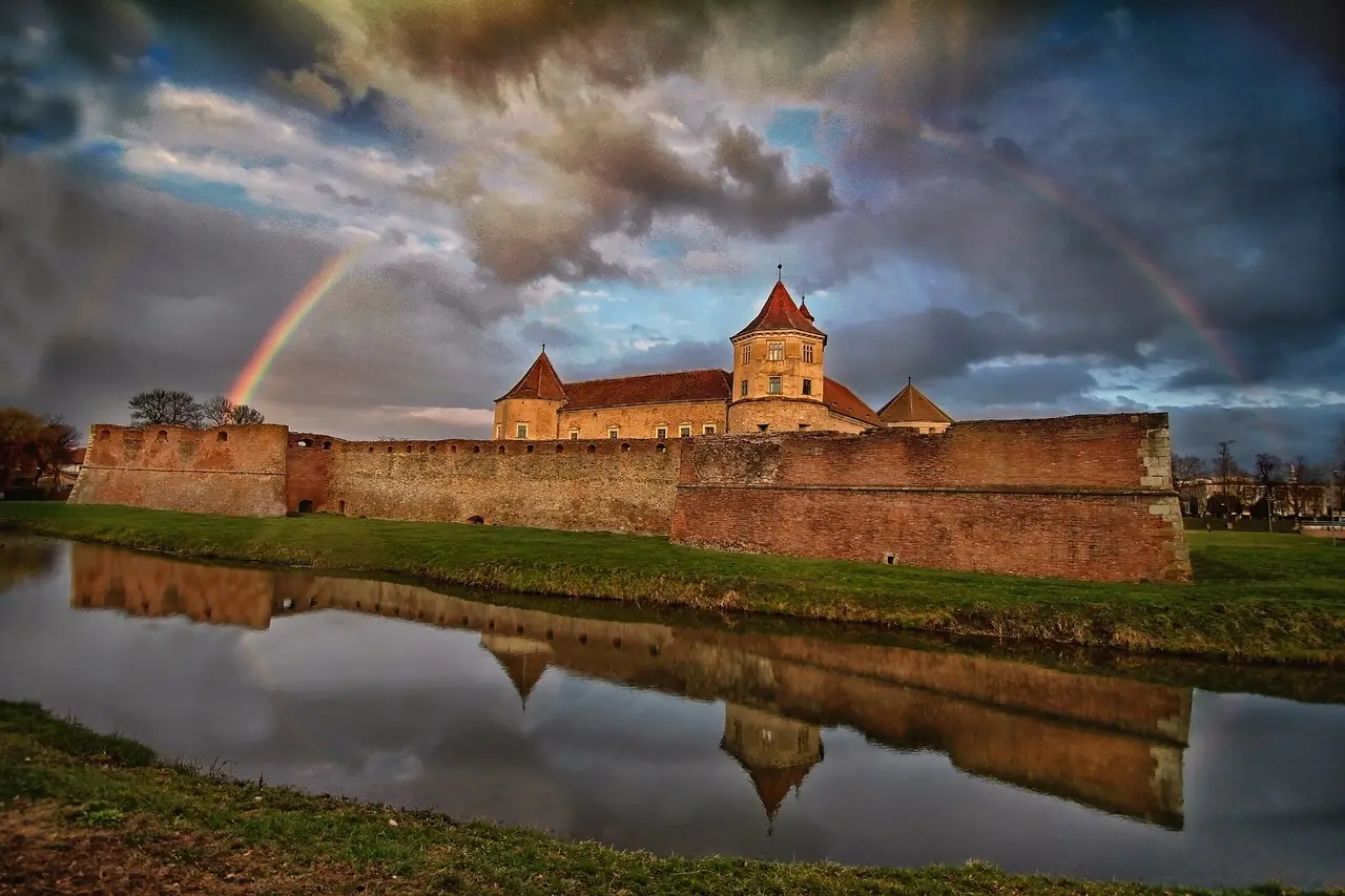 Fagaras citadel reflected in its moat under a dark sky, with a rainbow over the brick walls and red-tiled towers