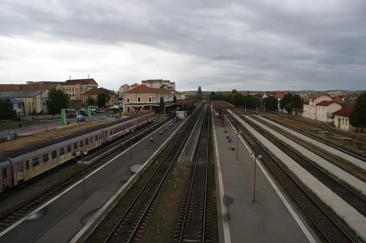Sibiu railway station seen from above, with a maroon CFR train at the platform, the historic station building and buses in the forecourt