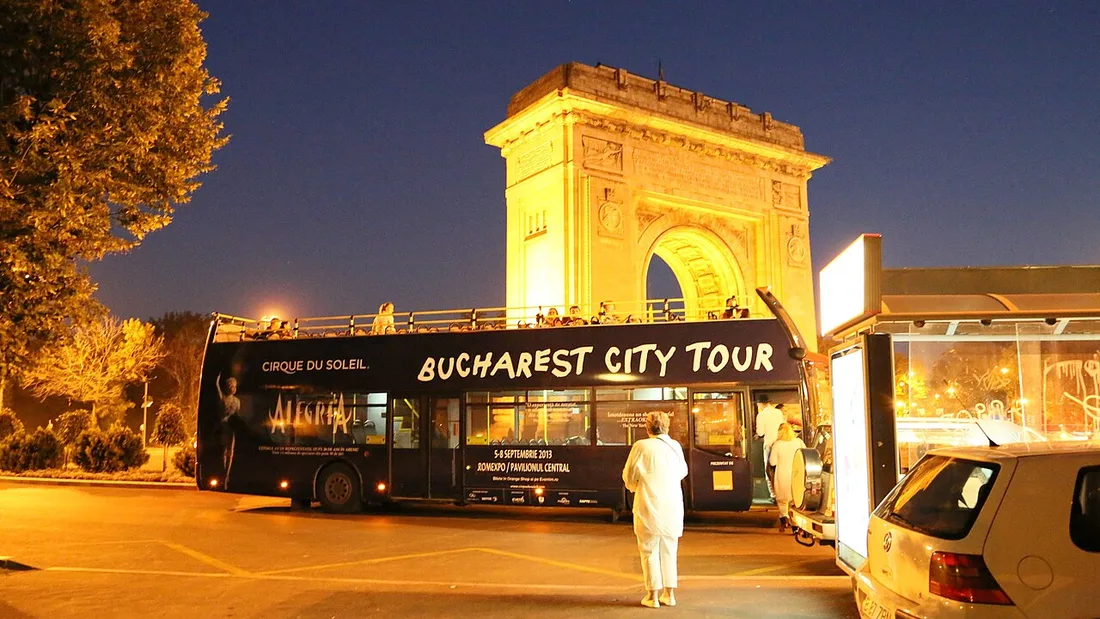The floodlit Arcul de Triumf on Kiseleff Road at dusk with a city sightseeing bus in front