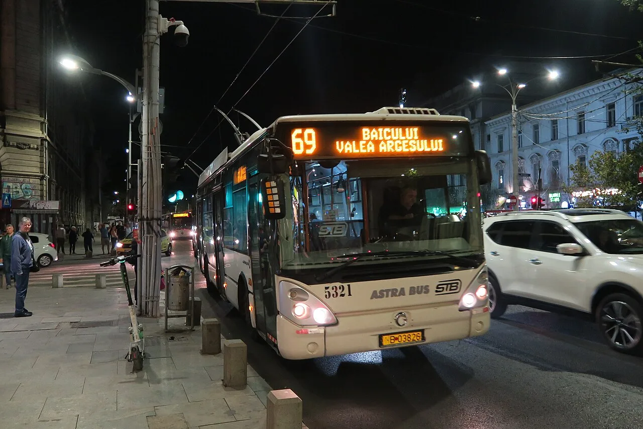 An STB city bus lit up at a stop on a central Bucharest street at night, its destination sign glowing orange, passers-by on the pavement