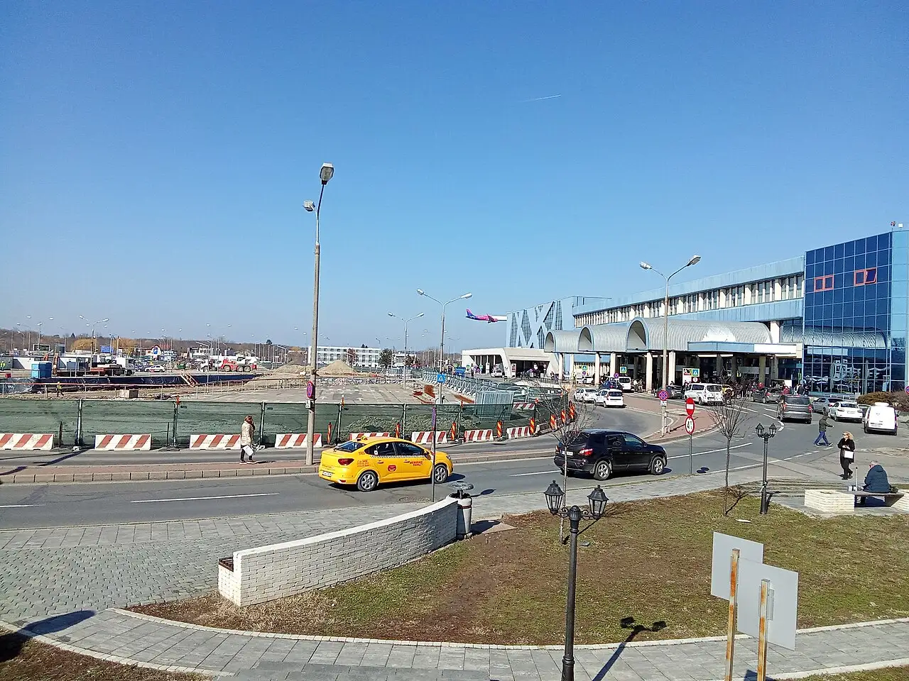 The curbside outside OTP terminal on a clear day, a bright yellow Bucharest taxi at the kerb, cars dropping off and a Wizz Air plane visible behind the terminal