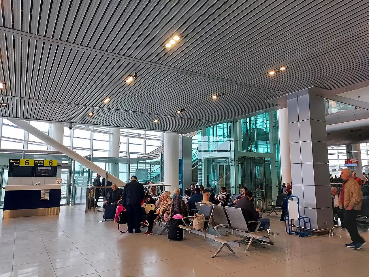 Inside the OTP terminal at Bucharest airport, travellers with luggage waiting near Gate 6 under a glass wall and a slatted metal ceiling