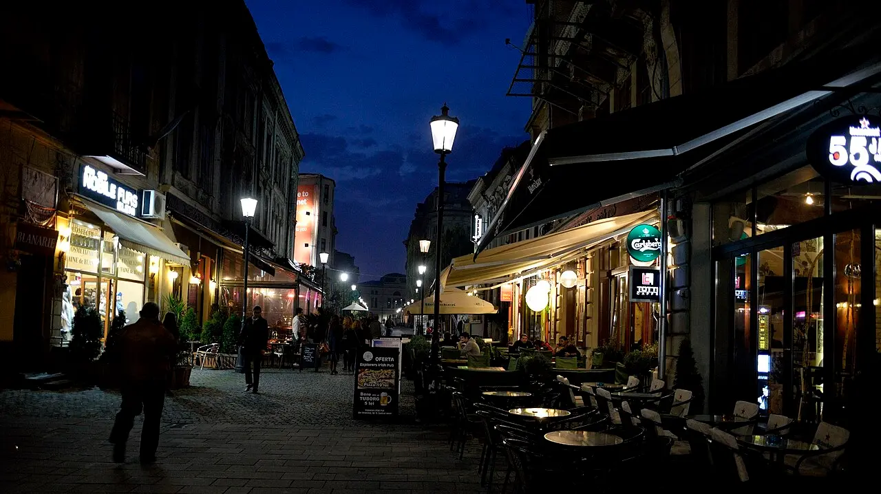 Bar terraces with awnings and street lamps on a cobbled Old Town lane in Bucharest at blue hour, people walking between the tables