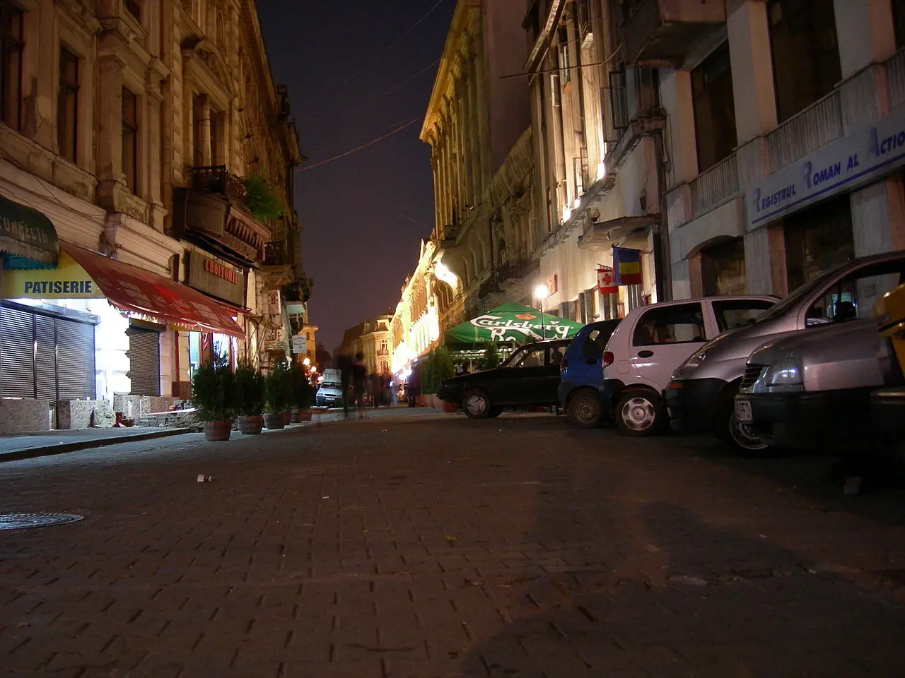 A pedestrian Old Town street in Bucharest at night with a lit patisserie, a Carlsberg parasol over a terrace and parked cars along the cobbles