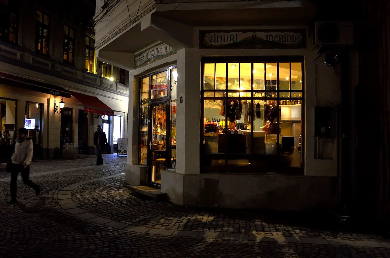 A warmly lit wine-and-deli shopfront on a quiet cobbled corner of the Bucharest Old Town at night, a couple walking past