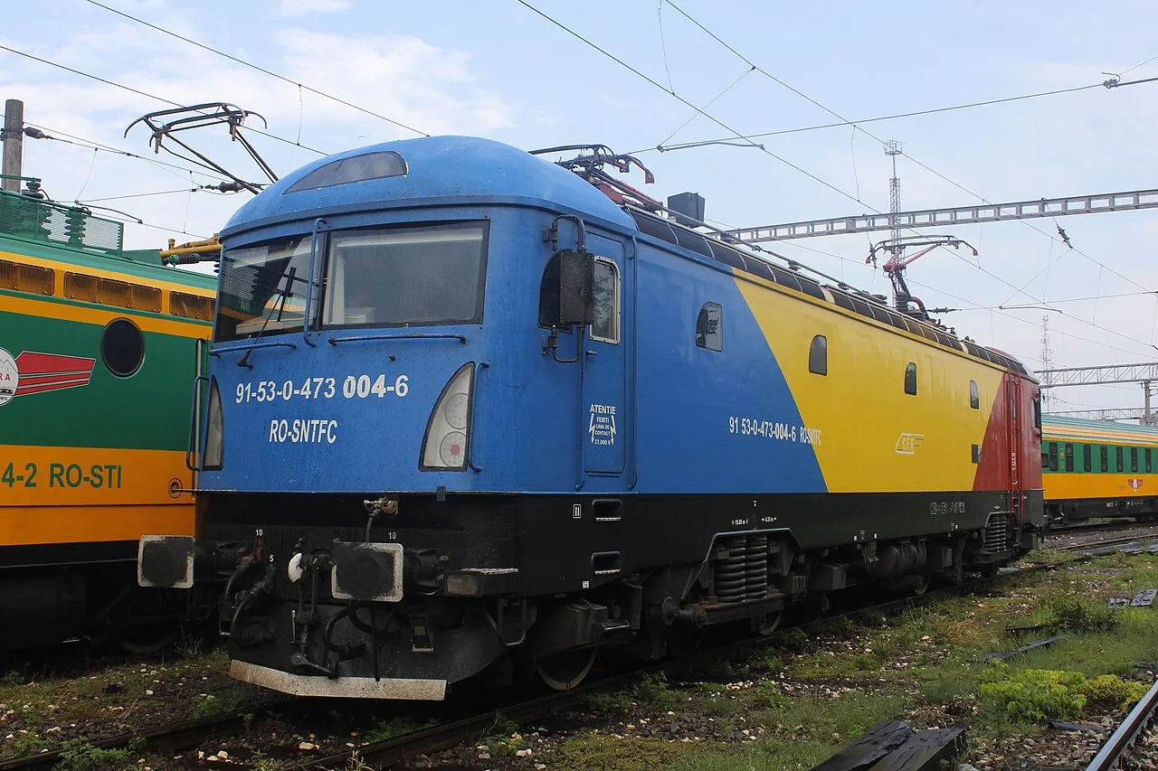 A CFR electric locomotive painted in the Romanian tricolour of blue, yellow and red standing in a rail yard
