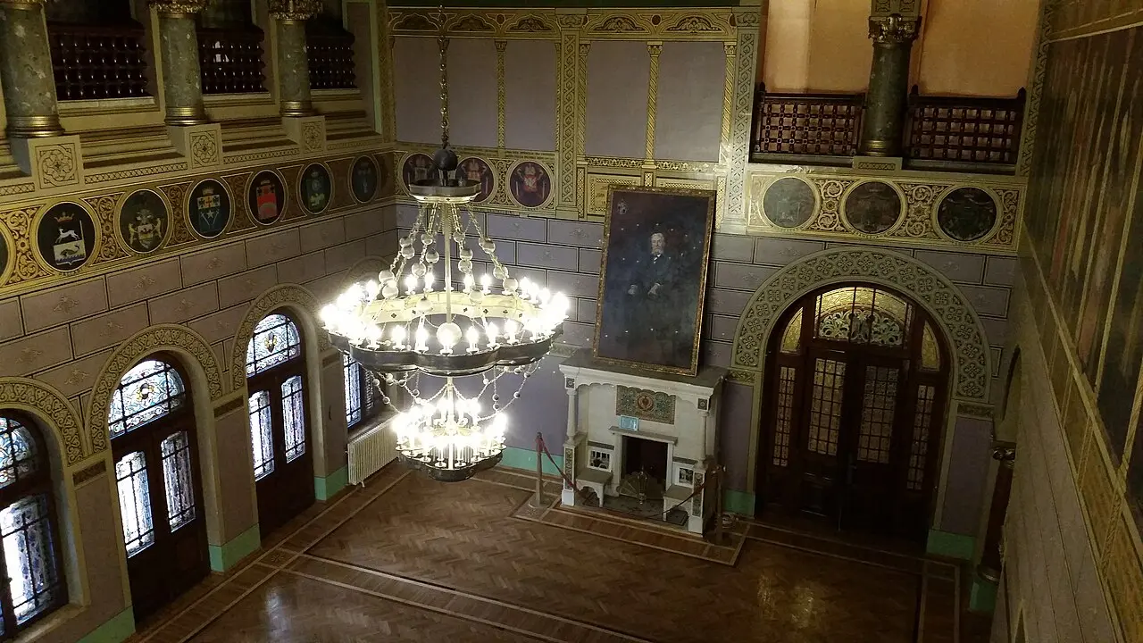 The ballroom of Cantacuzino Castle, with a large two-tier chandelier, a portrait of Prince Cantacuzino above a stone fireplace and heraldic medallions ringing the upper wall