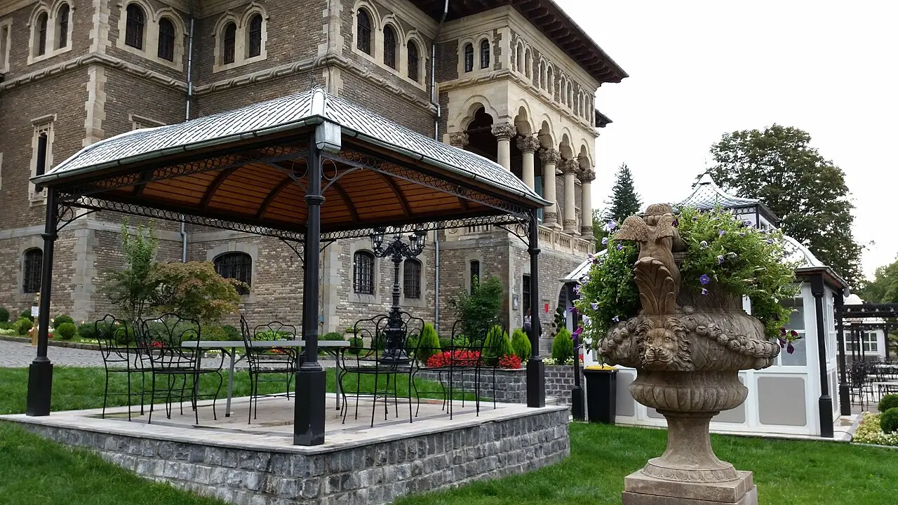 A wrought-iron pavilion and a carved stone urn planted with flowers in the courtyard of Cantacuzino Castle, with the arcaded loggia behind