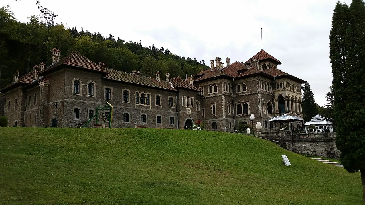 Cantacuzino Castle in Busteni seen from the front lawn, its grey stone wings and red-tiled towers backed by the forested slopes of the Bucegi mountains