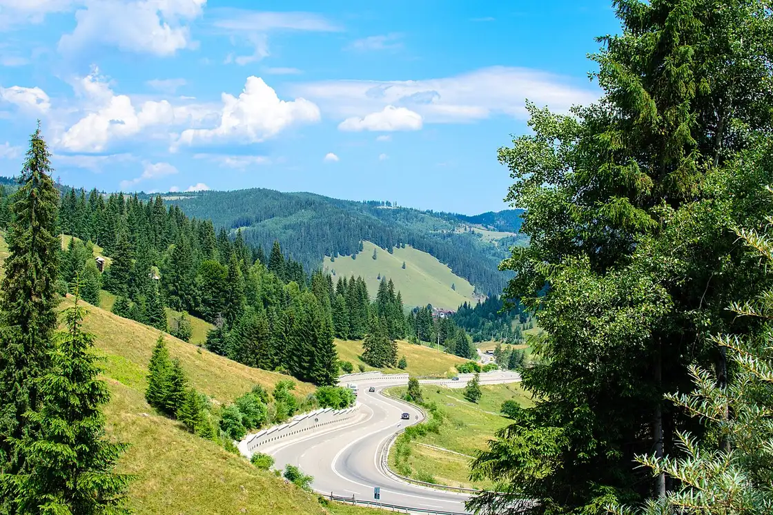 A winding two-lane road curving through green Carpathian hills under a blue summer sky, with a couple of cars on it and forested ridges behind