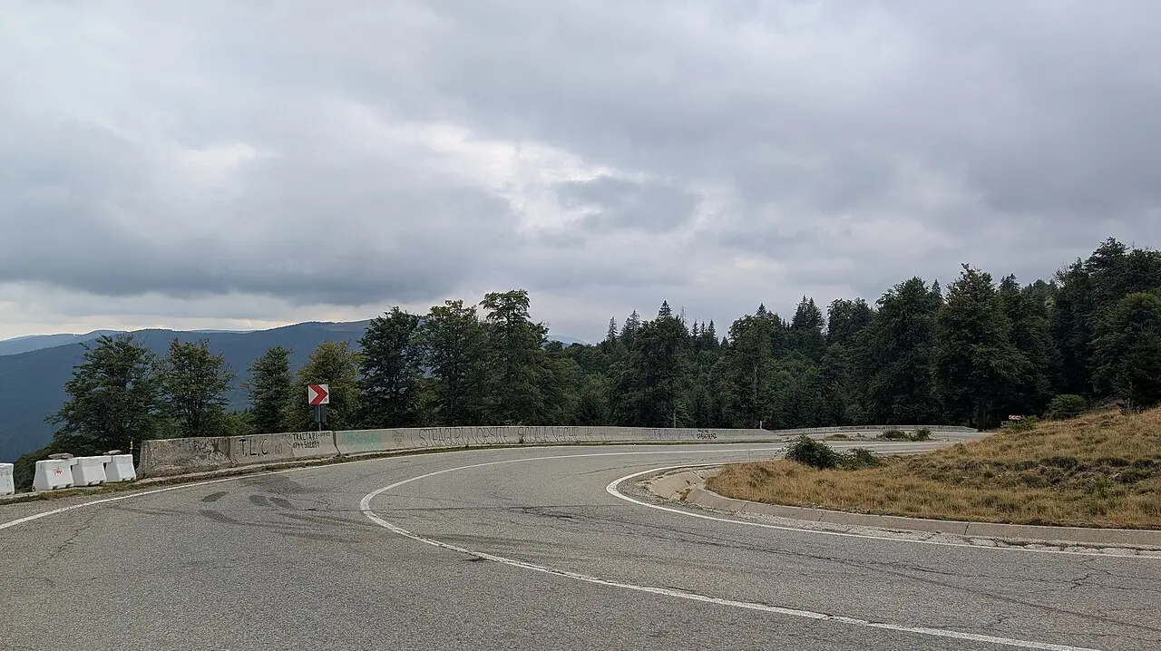 A hairpin bend on the Transalpina mountain road in Romania, the asphalt curving back on itself between forested Carpathian slopes under a grey sky