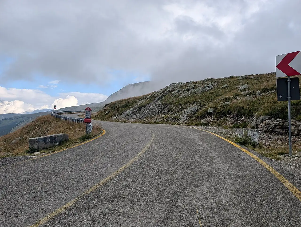 The Transalpina road high above the treeline in Romania, bare grassy ridges and a chevron warning sign, cloud rolling over the summit