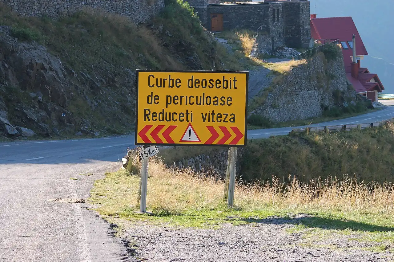 A yellow Romanian road sign on the Transfagarasan reading Curbe deosebit de periculoase, Reduceti viteza, warning of especially dangerous curves and a 15 km/h limit, with the road looping behind