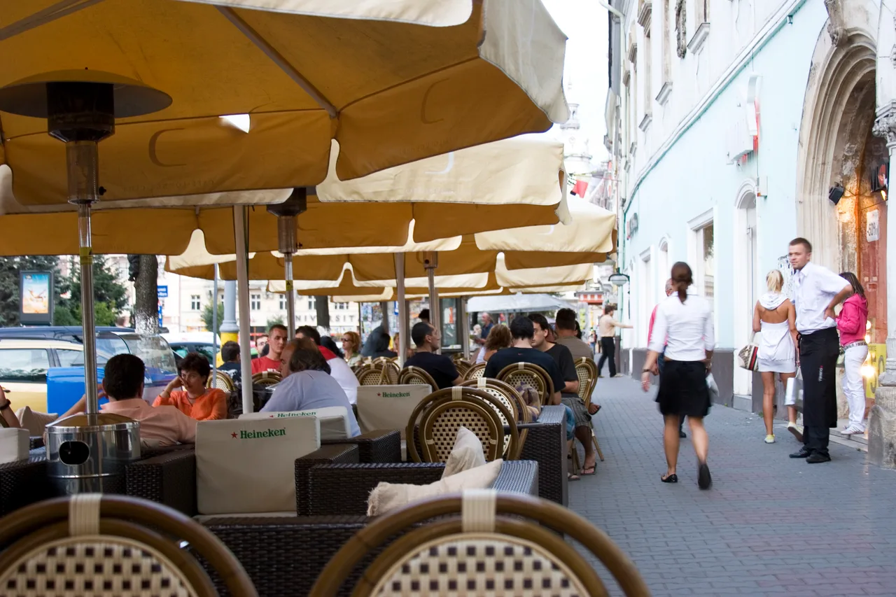 Busy cafe terraces under cream awnings on a pedestrian street in central Cluj-Napoca, people walking past in summer