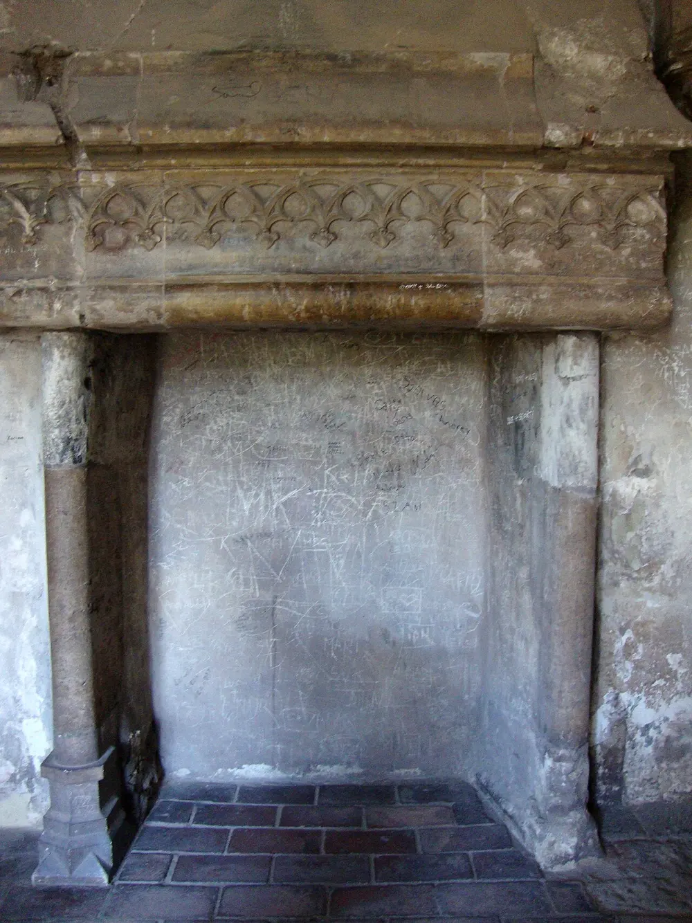 A carved stone fireplace inside Corvin Castle with Gothic tracery above and centuries of scratched graffiti on the wall behind it