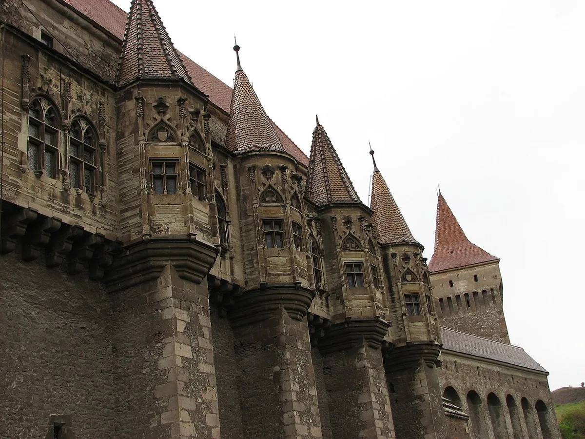 The Gothic oriel windows, carved stone balconies and pointed tiled spires along the side wall of Corvin Castle under a grey sky