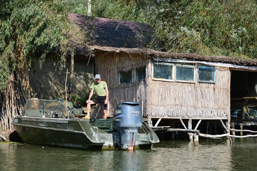 A fisherman standing in a small motorboat beside a reed-thatched stilt house on a Danube Delta channel