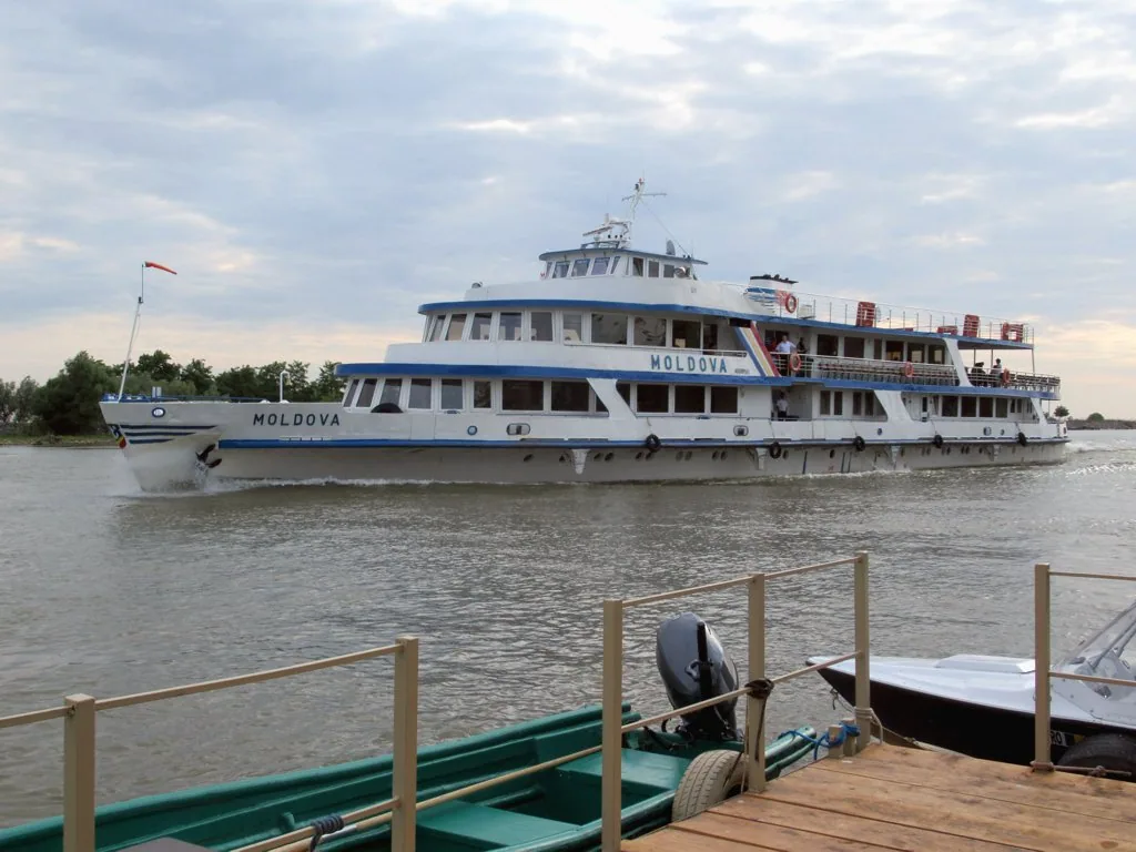 The Navrom passenger ferry Moldova on the Danube near a delta pier, with a traditional wooden boat moored in front