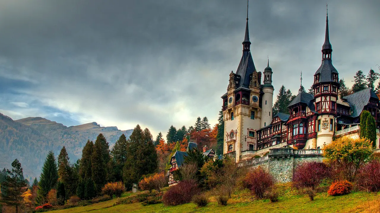 Peles Castle in Sinaia under autumn cloud, its turrets rising above pine forest and the Bucegi mountains