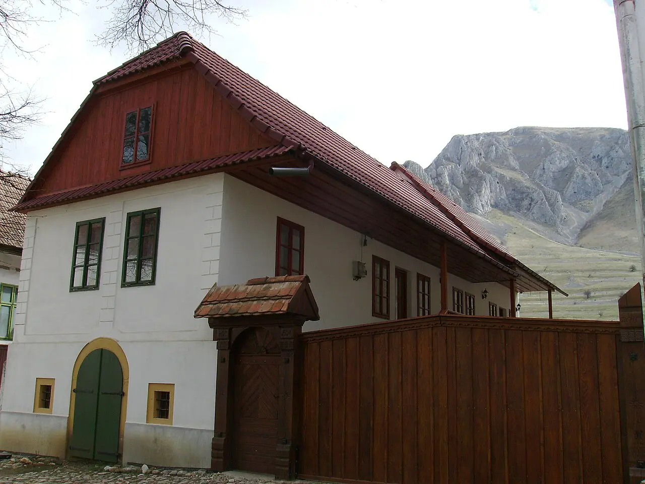 A white house with a green arched gate and red gable in Rimetea, with the limestone ridge of Piatra Secuiului rising behind