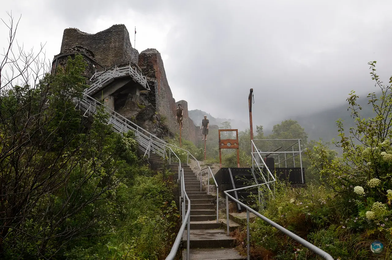The broken walls of Poenari Castle on a misty ridge, reached by a metal staircase, with impalement stakes re-created on the slope below