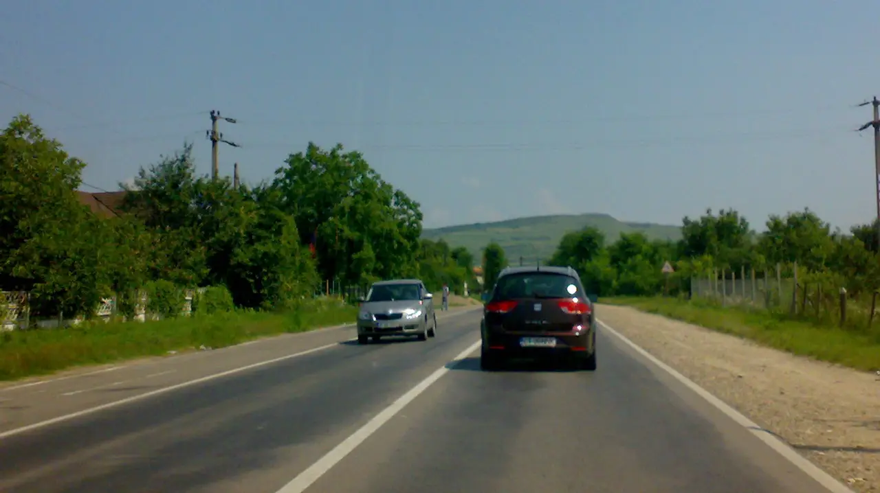 Two cars on a two-lane national road through a Romanian village, both with headlights on in daylight, low green hills ahead
