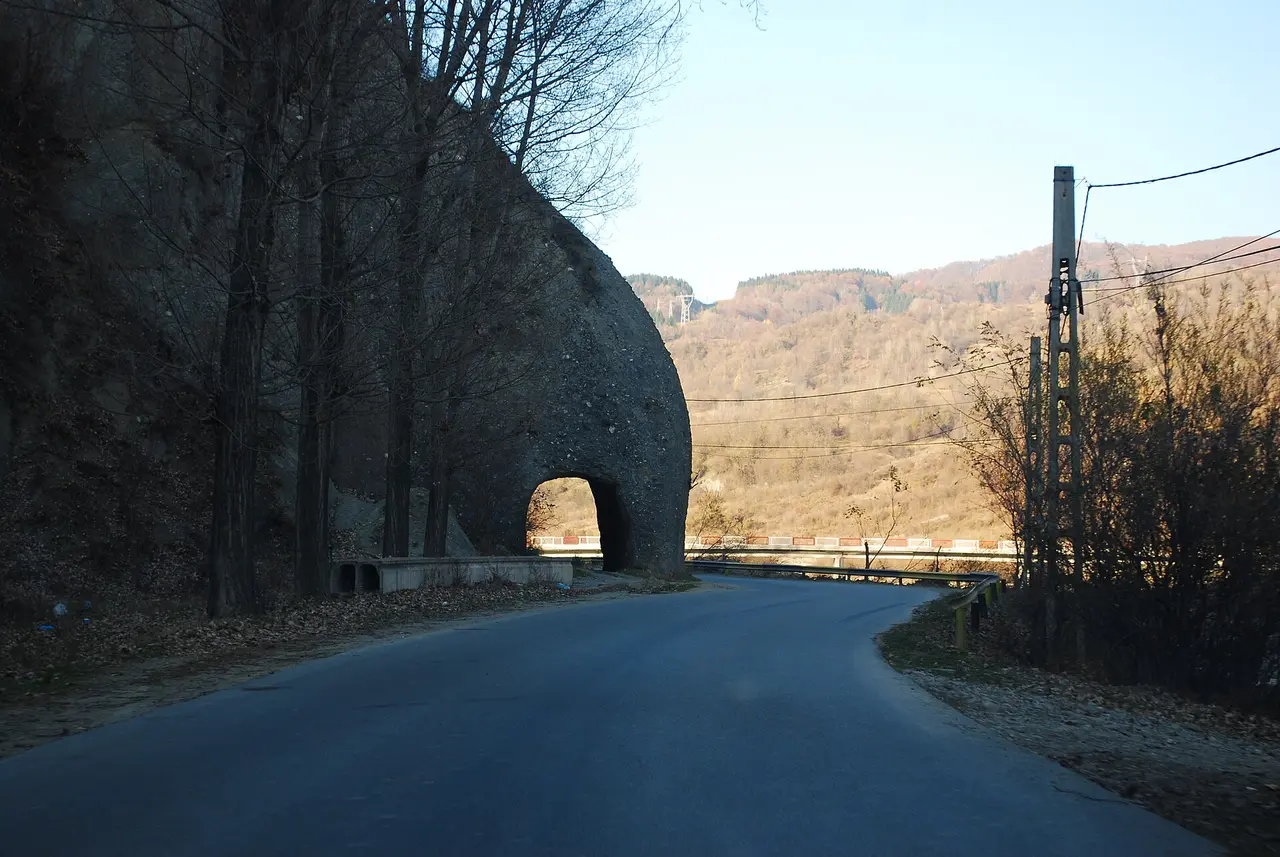 A narrow two-lane county road in the Doftana valley passing under a stone arch, forested hills behind and a crash barrier on the bend