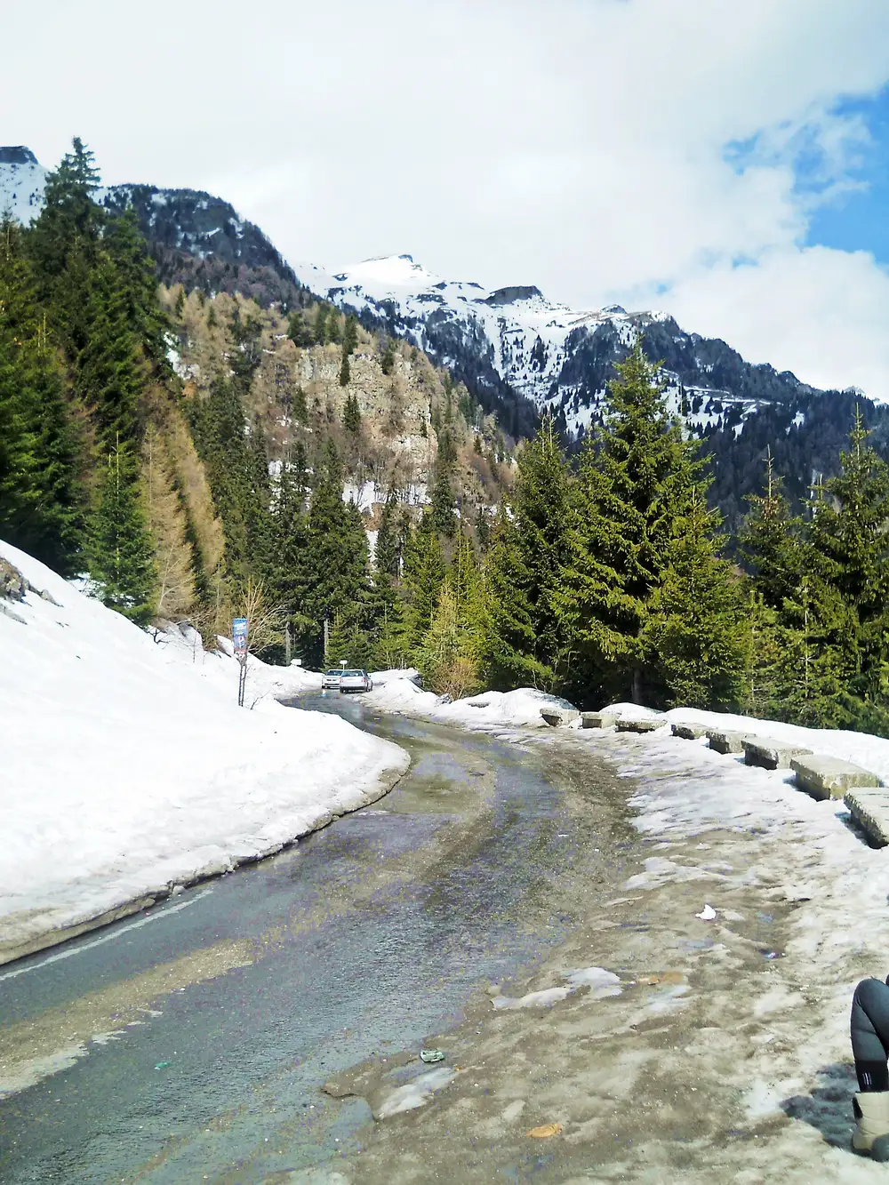 A mountain road in the Romanian Carpathians hemmed in by banks of melting snow, wet asphalt curving away and a car in the distance below snow-capped peaks
