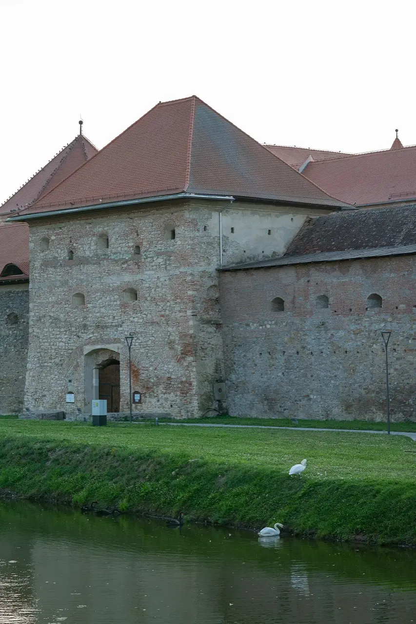 A brick bastion of Fagaras citadel rising from the moat, with swans on the water and autumn trees behind the walls