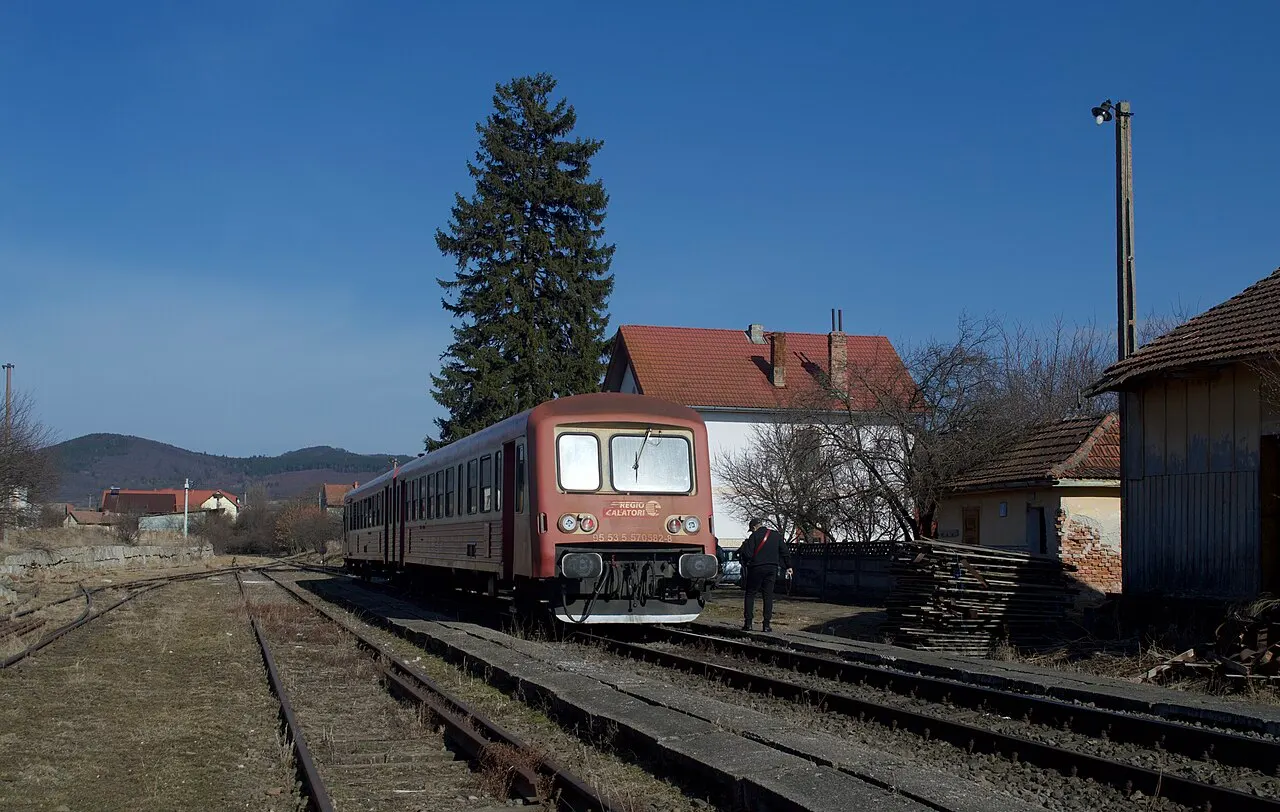 A blue and white CFR passenger train on a single-track rural line in the Eastern Carpathians, green hills behind