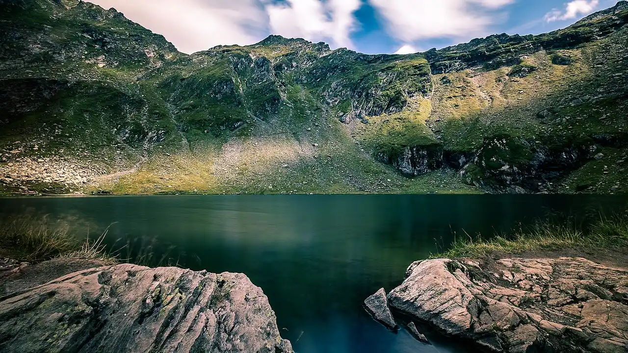 Balea Lake high in the Fagaras mountains, still dark water below steep green and grey slopes, boulders in the foreground under a bright sky