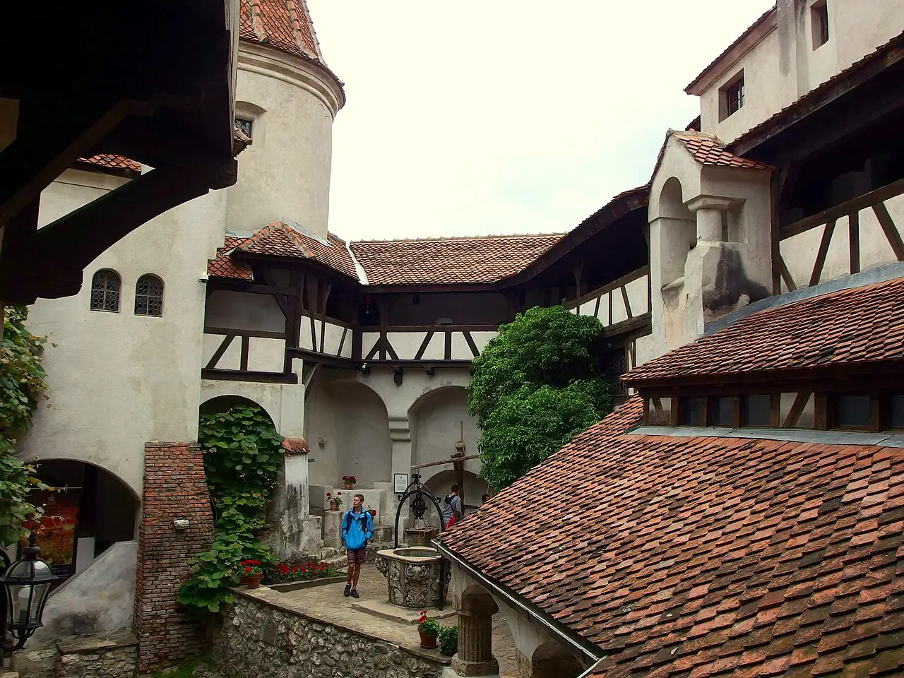 The inner courtyard of Bran Castle with its whitewashed walls, red-tiled roofs, timber galleries and the old well, a visitor in a blue jacket beside it
