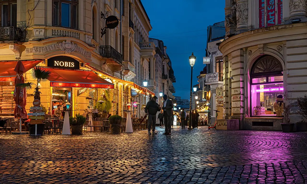 Cobblestone Lipscani street in the Old Town of Bucharest at blue hour, cafe terraces lit with fairy lights and a wet, glowing pavement