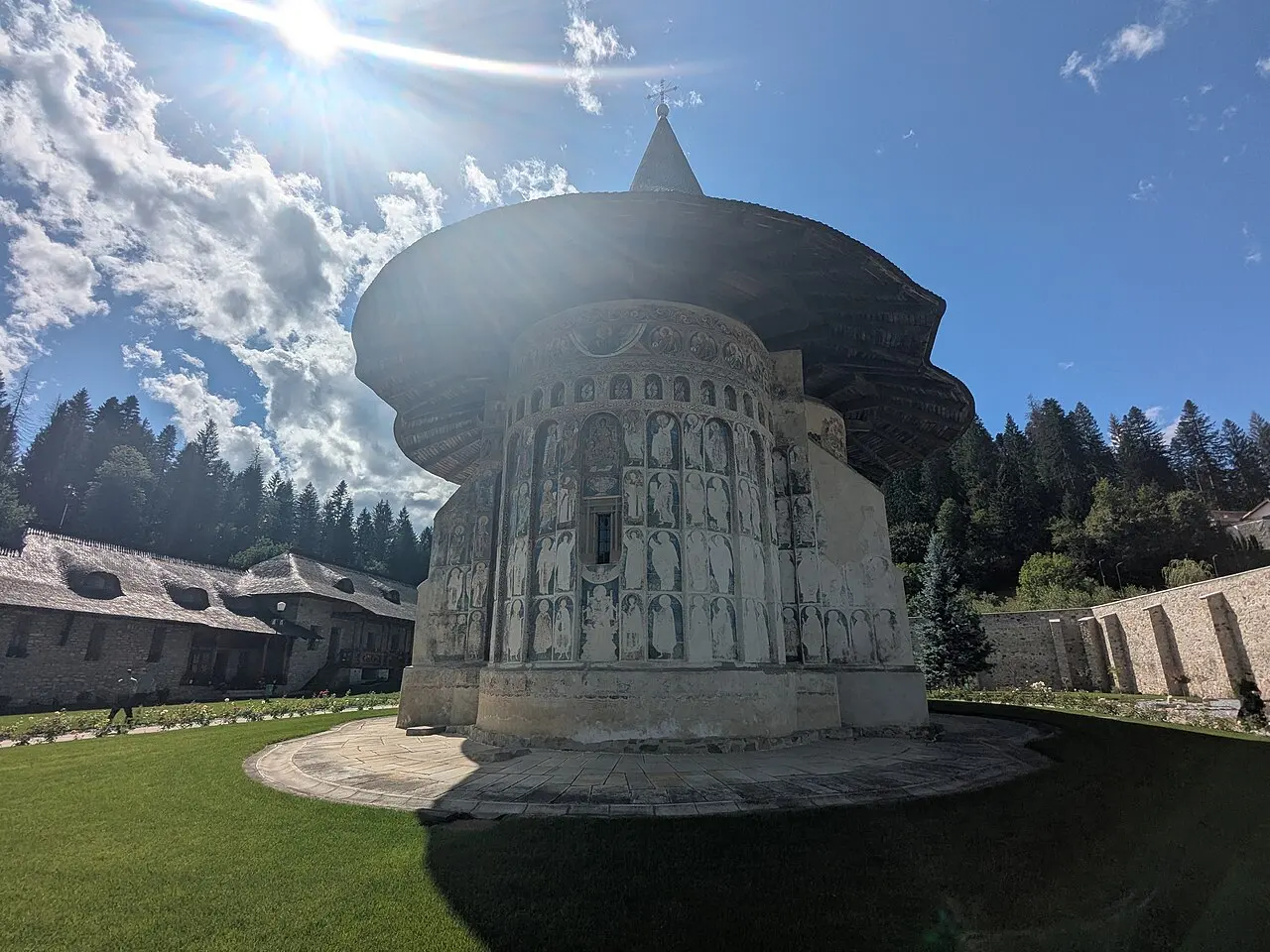 The frescoed exterior wall of Voronet Monastery in Bucovina, rows of painted saints under a shingled roof, green lawn and dark pines behind