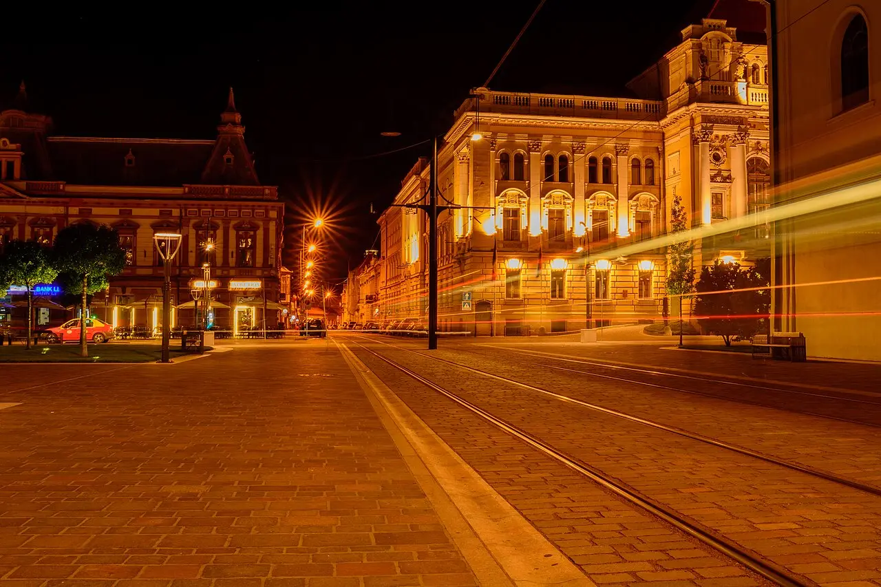 Union Square in Oradea lit up at night, the illuminated facades of the Moon Church and surrounding Secession buildings reflected on wet cobbles