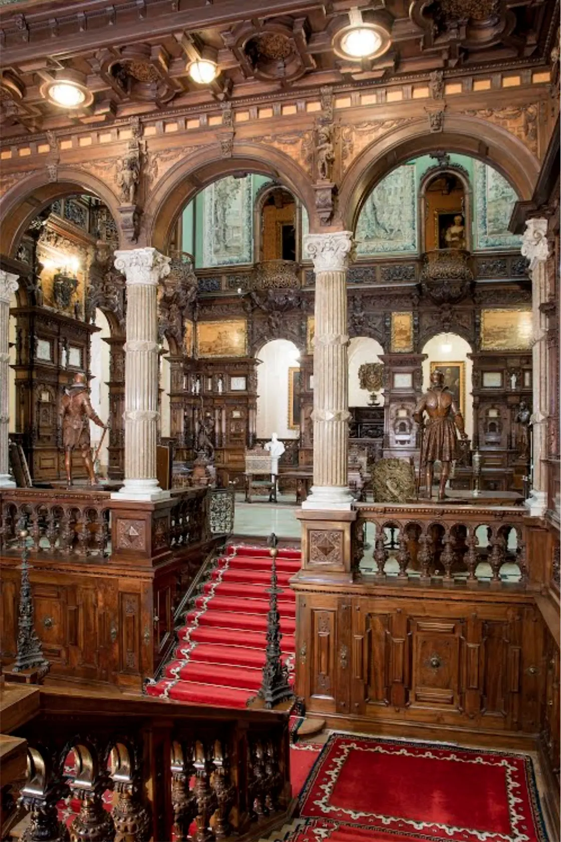 The Hall of Honour inside Peles Castle, walls covered in intricately carved walnut panelling rising several storeys to a stained-glass ceiling