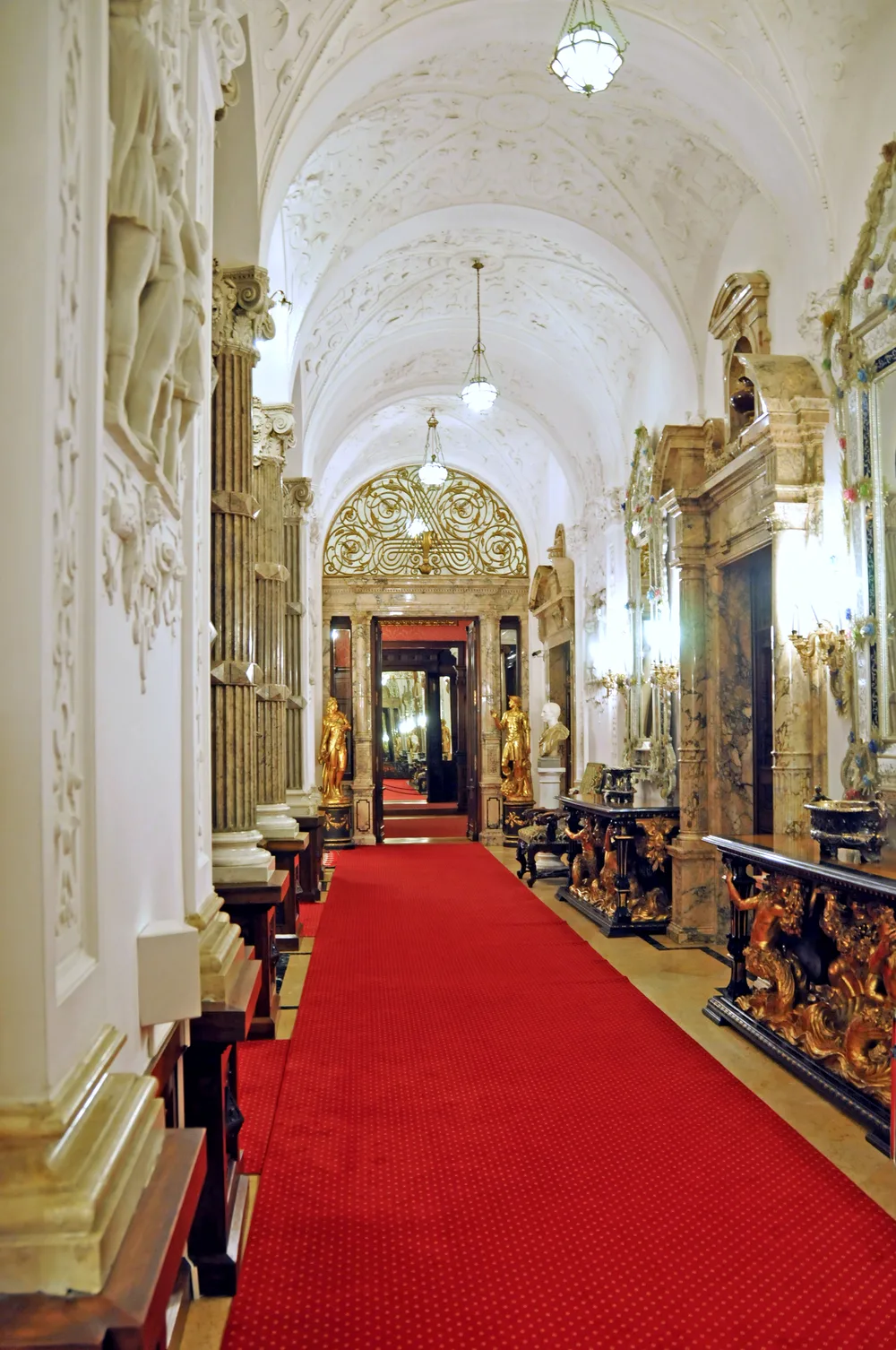 A narrow mirrored corridor inside Peles Castle, gilded and lit by chandeliers, reflections receding into the distance