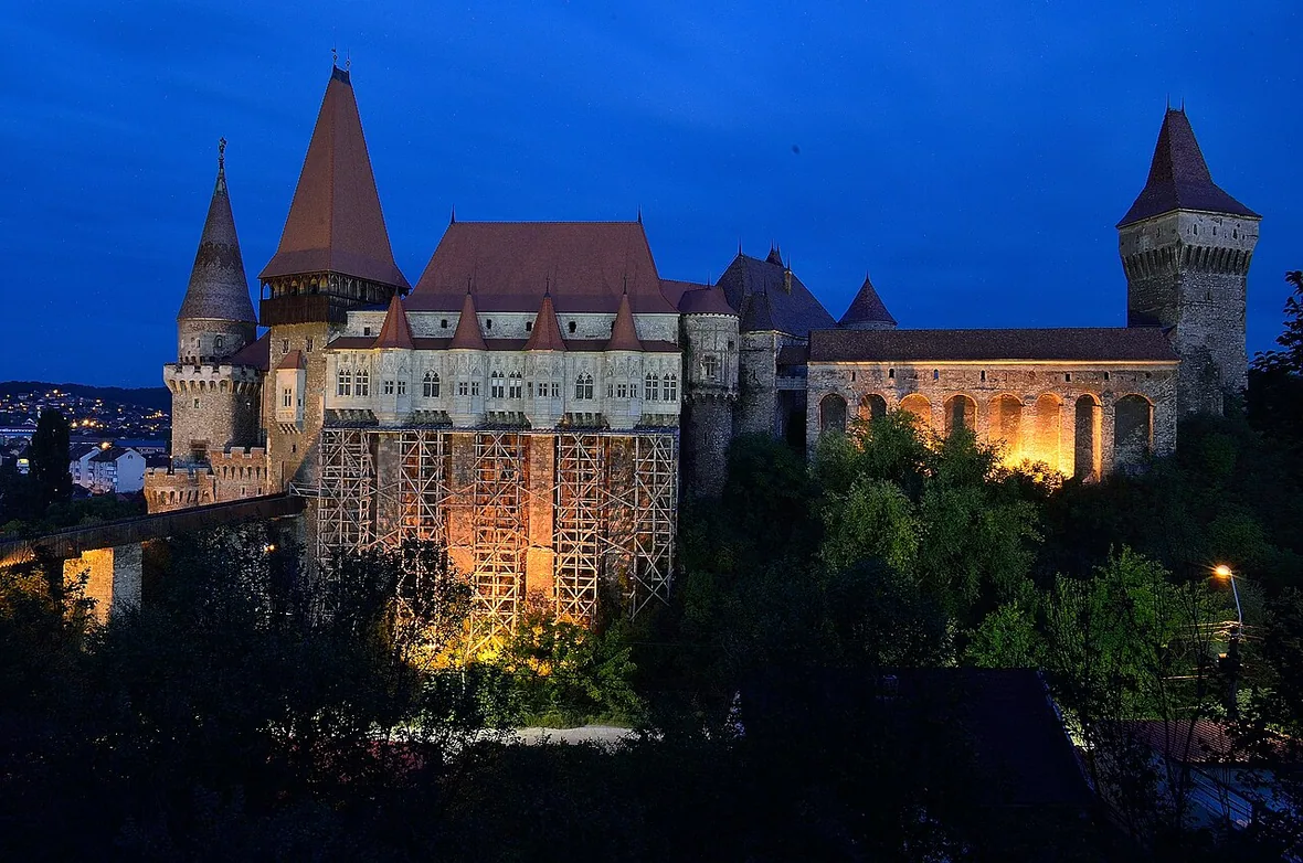 Corvin Castle floodlit at dusk, its towers and arcaded wing glowing against a deep blue sky