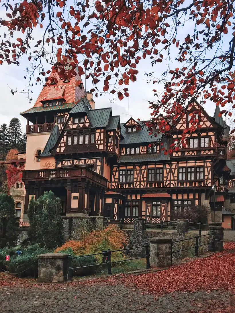 Pelisor Castle in late autumn, framed by red beech leaves, with fallen leaves covering the cobbled lane in front of the stone terrace